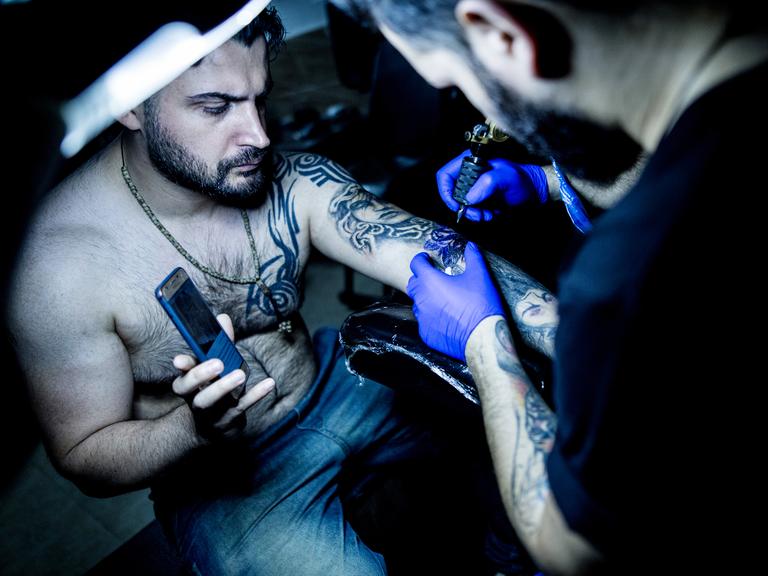 Ein iranischer Tätowierer in seinem Tattoostudio bei der Arbeit, er tätowiert einen Mann dessen Haut bei einem Unfall verbrannt wurde, Gilan im Iran, 2019.