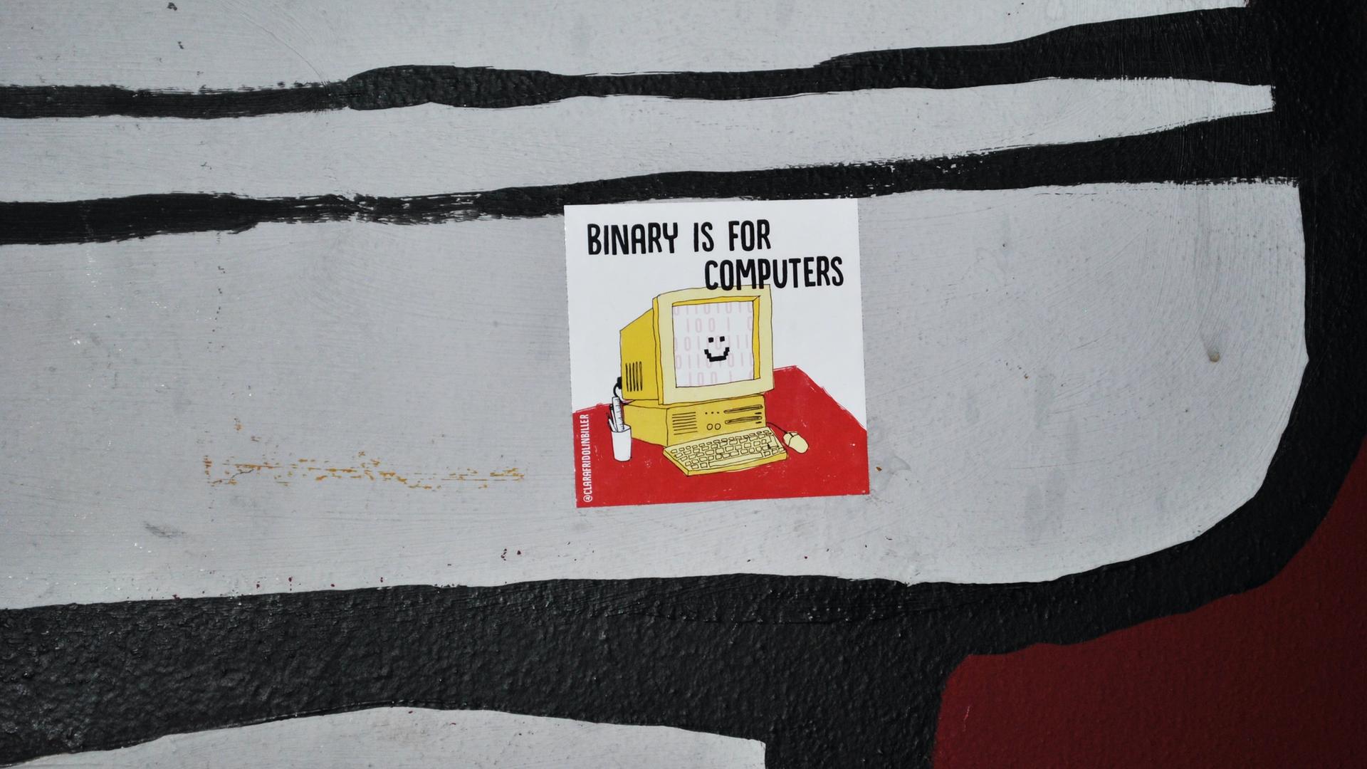 Auf einer Oberfläche klebt ein Sticker mit der Illustration eines lächelnden Computers und der Aufschrift "Binary is for Computers".