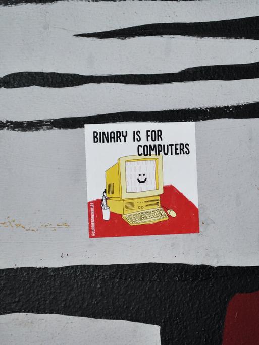 Auf einer Oberfläche klebt ein Sticker mit der Illustration eines lächelnden Computers und der Aufschrift "Binary is for Computers".