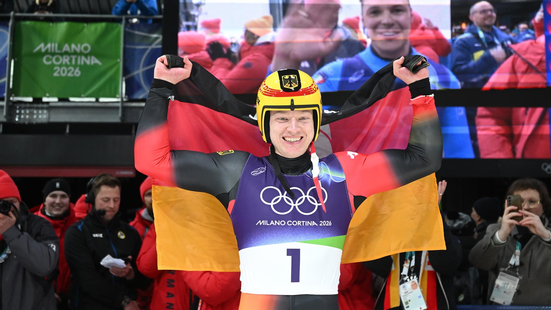 Max Langenhan trägt einen Rodleranzug und einen gelben Helm, er jubelt mit einer Deutschlandflagge über den Schultern.