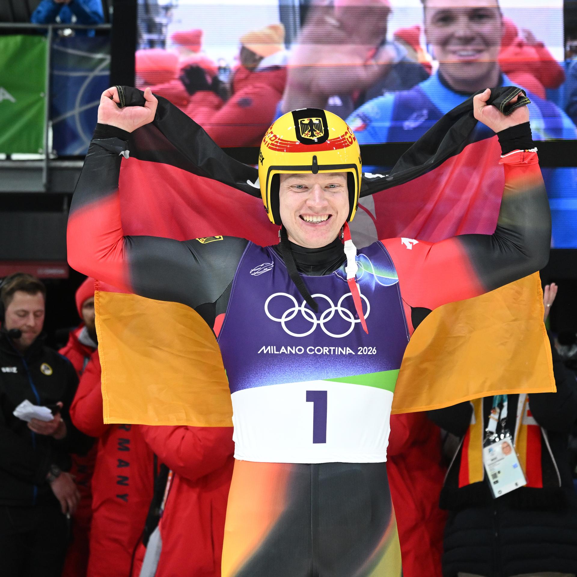 Max Langenhan trägt einen Rodleranzug und einen gelben Helm, er jubelt mit einer Deutschlandflagge über den Schultern.