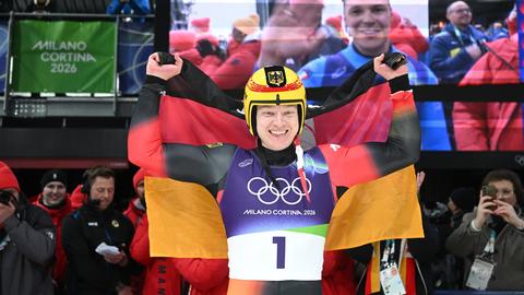 Max Langenhan trägt einen Rodleranzug und einen gelben Helm, er jubelt mit einer Deutschlandflagge über den Schultern.
