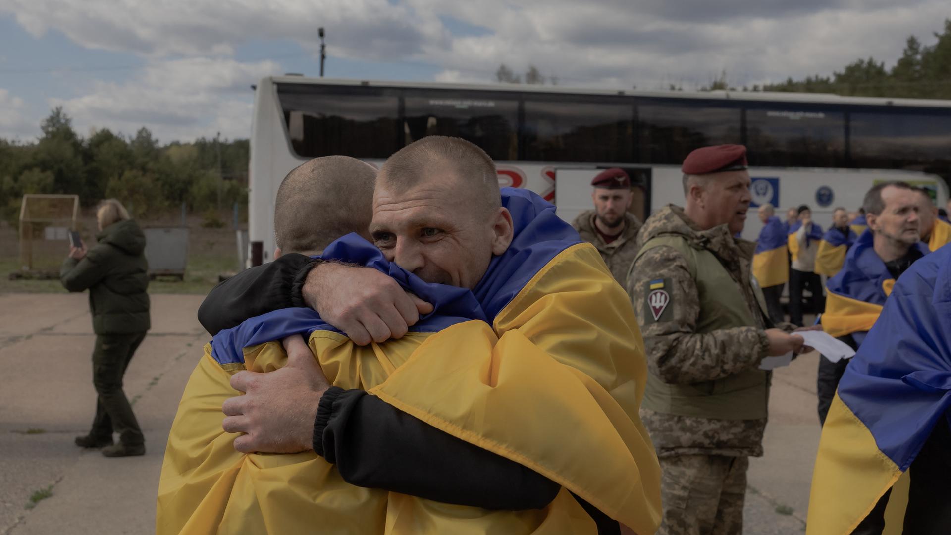 Zwei Männer liegen sich in den Armen. Sie tragen ukrainische Flaggen über den Schultern und stehen vor einem Bus. Im Hintergrund stehen weitere Menschen mit ukrainischen Flaggen sowie ein Soldat. 