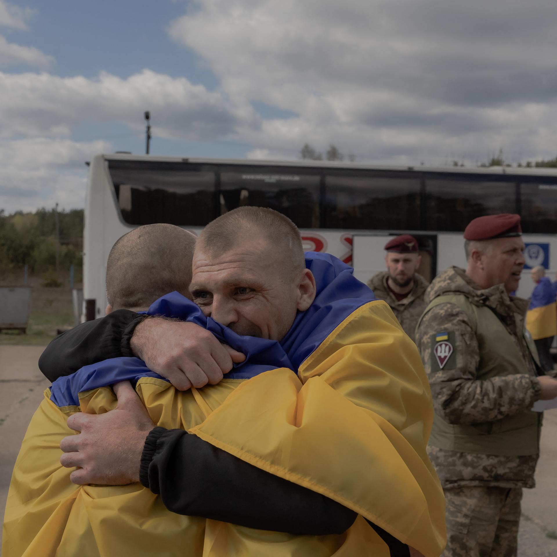 Zwei Männer liegen sich in den Armen. Sie tragen ukrainische Flaggen über den Schultern und stehen vor einem Bus. Im Hintergrund stehen weitere Menschen mit ukrainischen Flaggen sowie ein Soldat. 