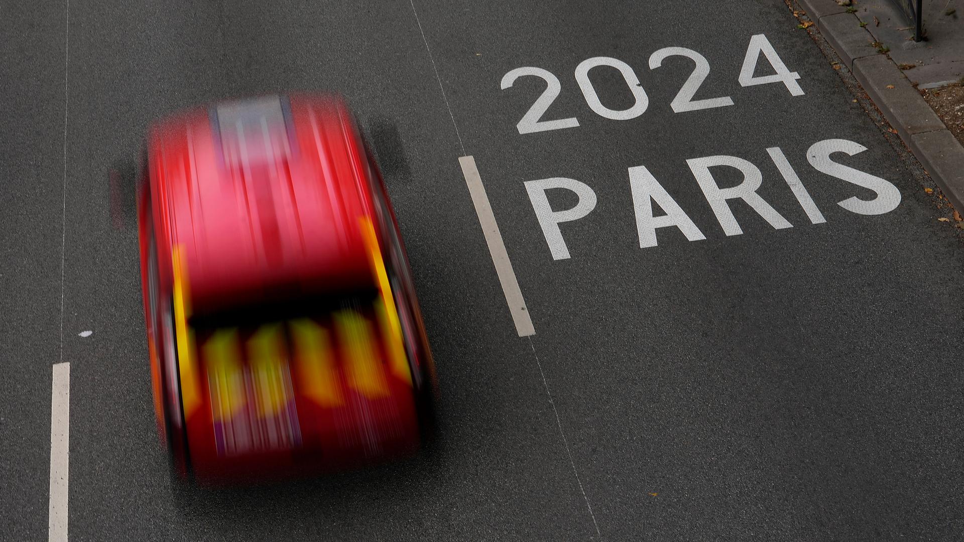 Ein rotes Taxi fährt über eine Straße. Auf der Fahrbahn steht 2024 Paris.