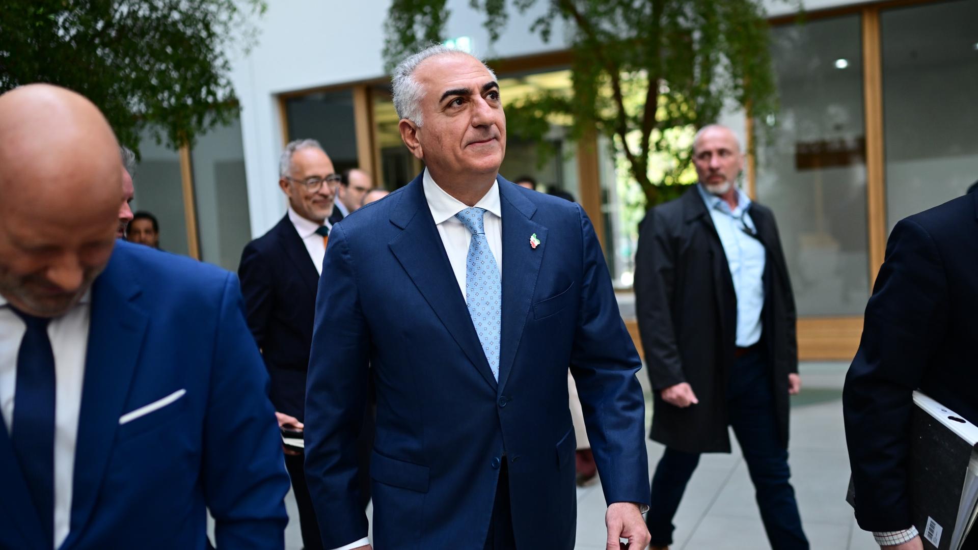 Reza Pahlavi kommt zur Bundespressekonferenz. Er wird von privaten Sicherheitskräften begleitet. Er trägt einen blauen Anzug und eine hellblaue Krawatte.