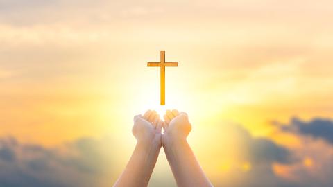 Zwei als Schale geformte Hände zeigen Richtung Himmel, über den Händen schwebt ein Holzkreuz.