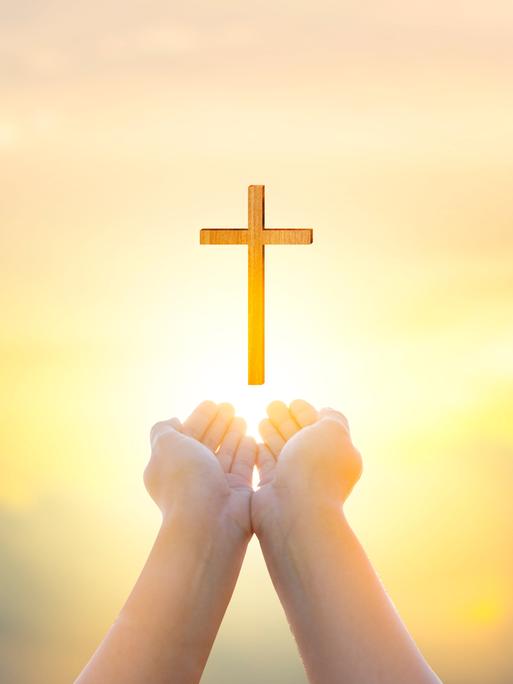 Zwei als Schale geformte Hände zeigen Richtung Himmel, über den Händen schwebt ein Holzkreuz.