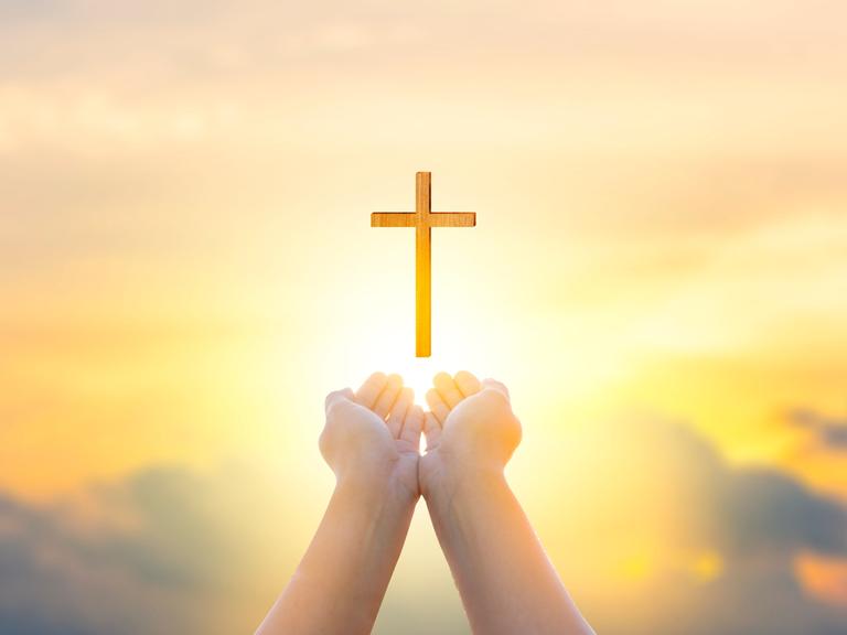 Zwei als Schale geformte Hände zeigen Richtung Himmel, über den Händen schwebt ein Holzkreuz.