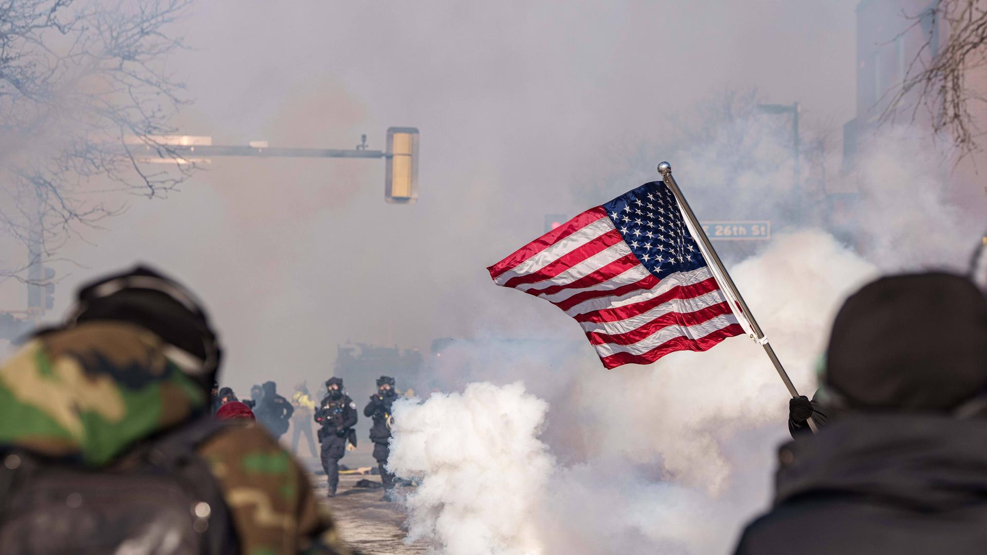 Rauch auf der Straße, Beamte von ICE und Demonstranten und im Vordergrund eine US-Flagge.