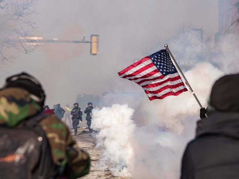 Rauch auf der Straße, Beamte von ICE und Demonstranten und im Vordergrund eine US-Flagge.