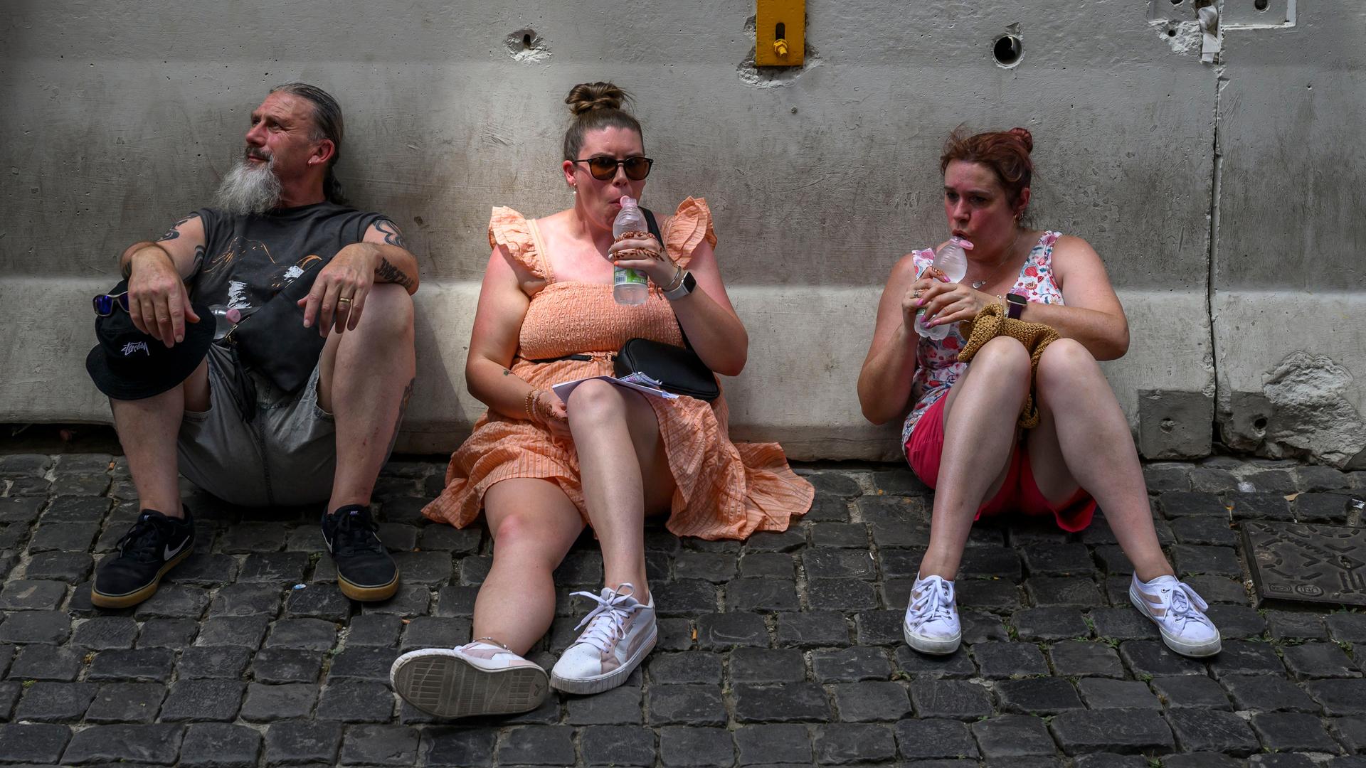 Drei Personen sitzend schwitzend und gerötet im Schatten auf dem Boden und trinken aus Wasserflaschen.