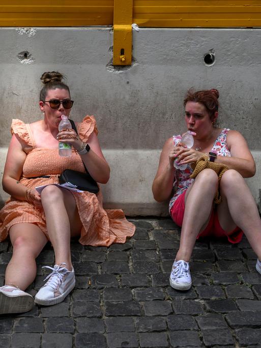 Drei Personen sitzend schwitzend und gerötet im Schatten auf dem Boden und trinken aus Wasserflaschen.