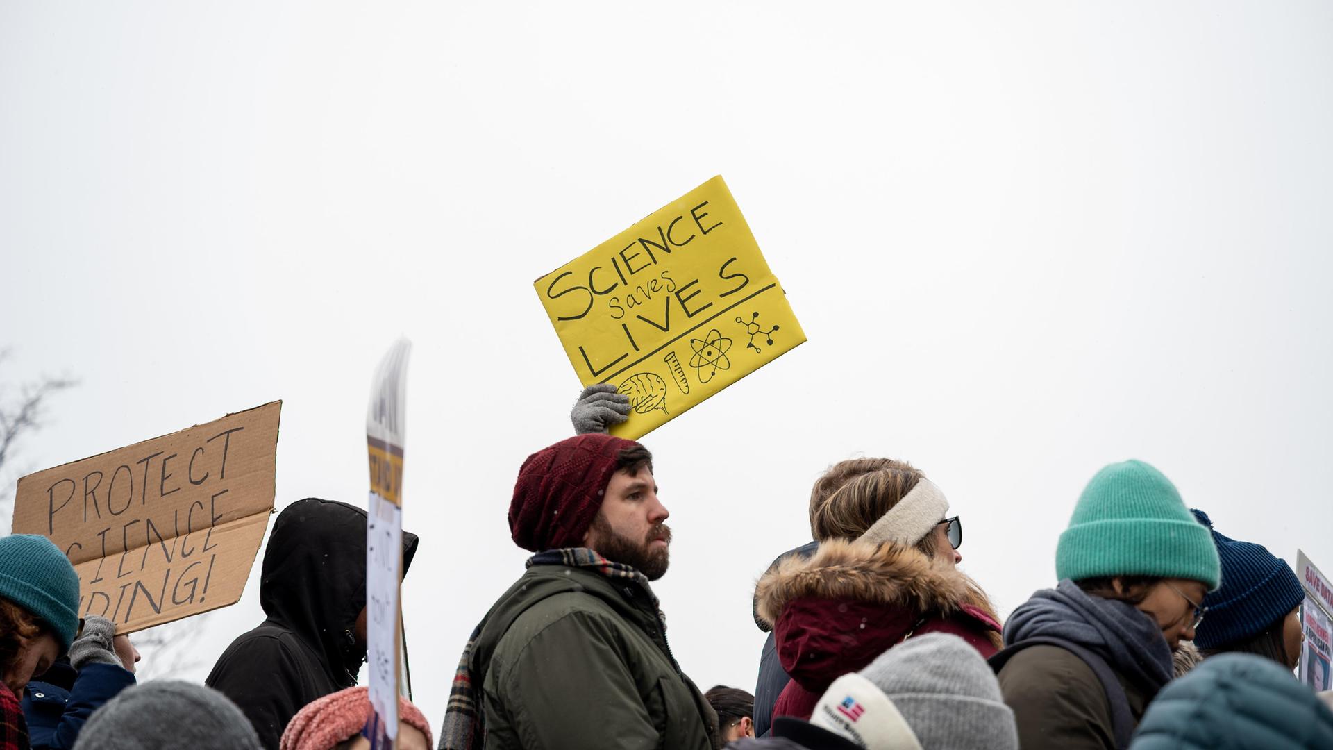 Protest von Wissenschaftlern, Forschern und ihren Unterstützern. Ein gelbes Schild wird in die Höhe gehalten. Darauf steht "Science saves lives", zu deutsch Wissenschaft rettet Leben.