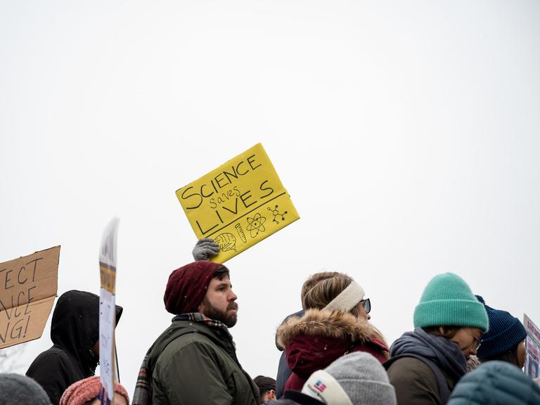 Protest von Wissenschaftlern, Forschern und ihren Unterstützern. Ein gelbes Schild wird in die Höhe gehalten. Darauf steht "Science saves lives", zu deutsch Wissenschaft rettet Leben.