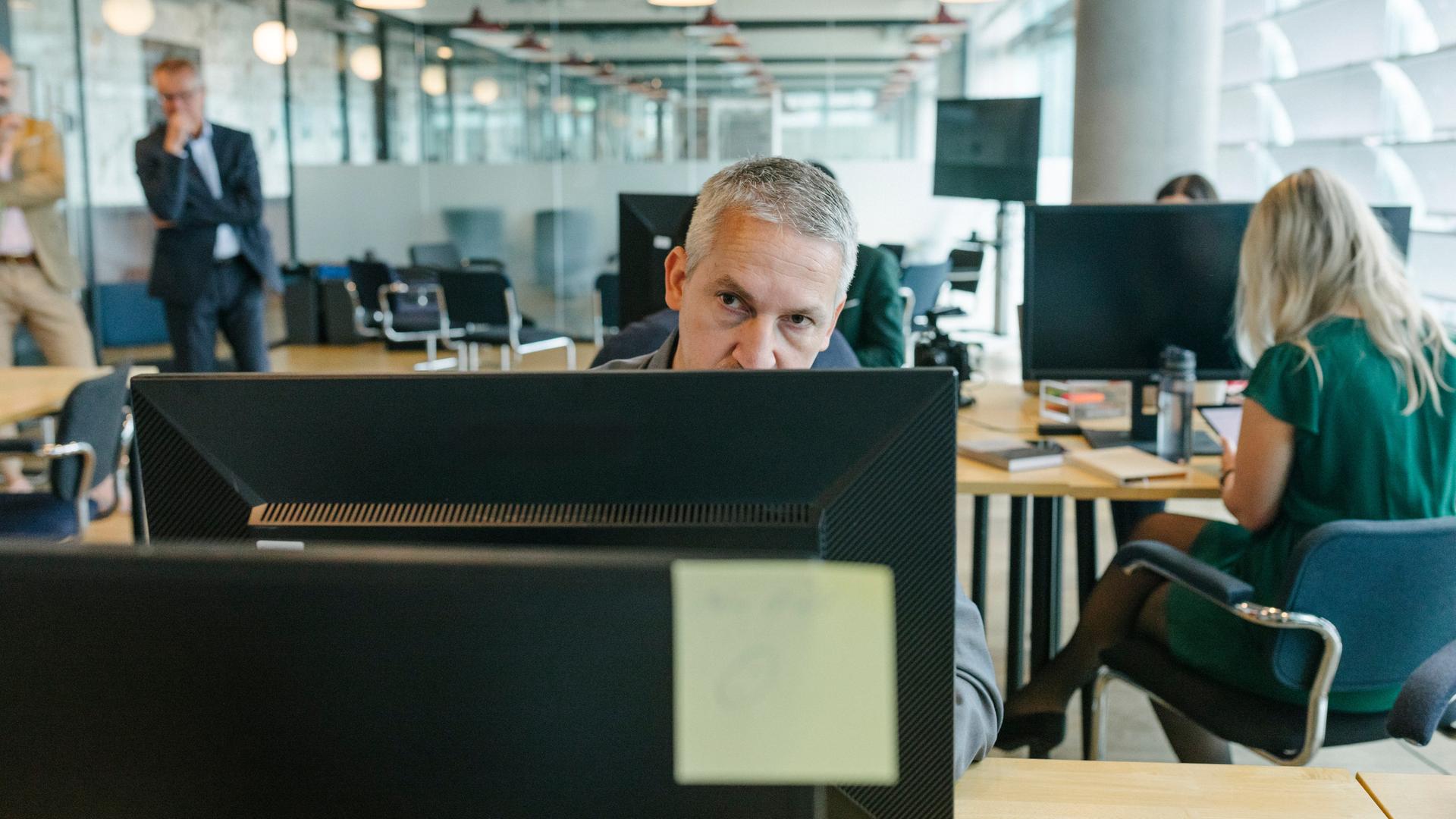 Ein Mann schaut mit schlecht gelauntem Blick über den Rand seines Monitors in einem Großraumbüro.