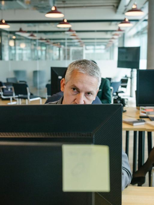 Ein Mann schaut mit schlecht gelauntem Blick über den Rand seines Monitors in einem Großraumbüro.