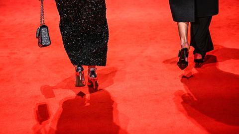 Zwei Frauen mit hochhackigen Schuhen gehen über einen roten Teppich. Nur die Füße von hinten sind abgebildet. Die linke Frau trägt ein schwarzes Glitzerkleid und eine Handtasche, die rechte einen schwarzen Rock, der an einem Bein zu einer Hose wird.