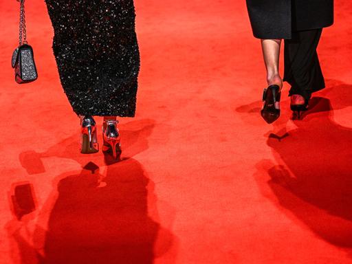Zwei Frauen mit hochhackigen Schuhen gehen über einen roten Teppich. Nur die Füße von hinten sind abgebildet. Die linke Frau trägt ein schwarzes Glitzerkleid und eine Handtasche, die rechte einen schwarzen Rock, der an einem Bein zu einer Hose wird.