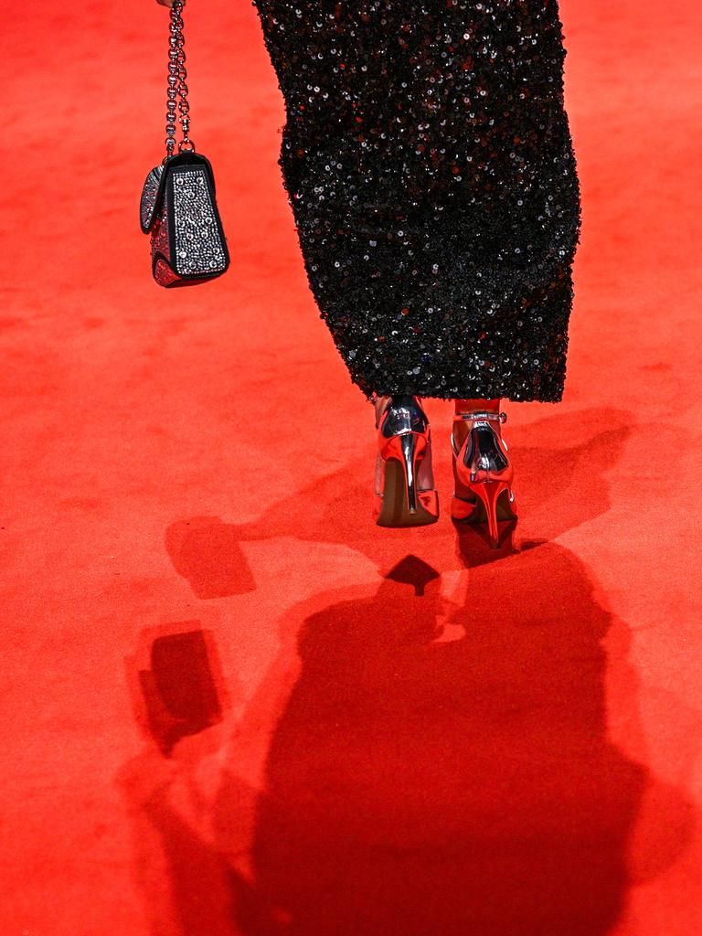 Zwei Frauen mit hochhackigen Schuhen gehen über einen roten Teppich. Nur die Füße von hinten sind abgebildet. Die linke Frau trägt ein schwarzes Glitzerkleid und eine Handtasche, die rechte einen schwarzen Rock, der an einem Bein zu einer Hose wird.