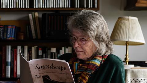 Eine Frau mit schulterlangen Haaren und Brille sitzt auf einem Sessel und liest in einer Partitur mit dem Titel "Soundscape"