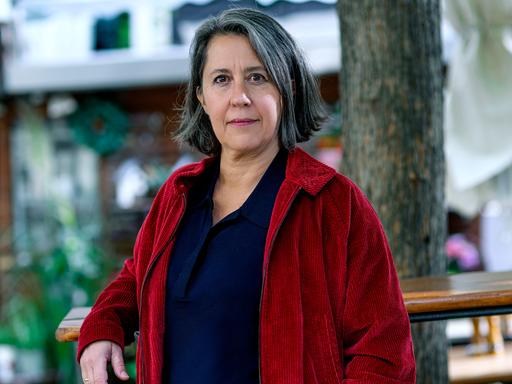 Eine Frau steht in einem Gastgarten und blickt ernst in die Kamera. Sie trägt Jeans, ein dunkelblaues Oberteil und eine rote Cordjacke. Ihre Haare sind grau bis dunkelbraun, ihre Augen braun.