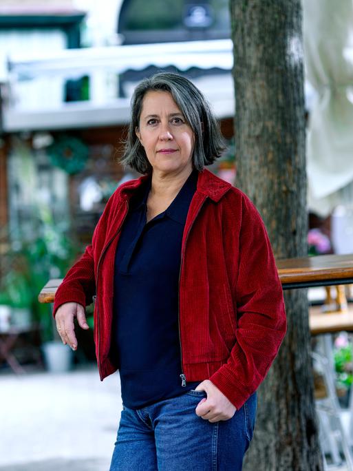 Eine Frau steht in einem Gastgarten und blickt ernst in die Kamera. Sie trägt Jeans, ein dunkelblaues Oberteil und eine rote Cordjacke. Ihre Haare sind grau bis dunkelbraun, ihre Augen braun.