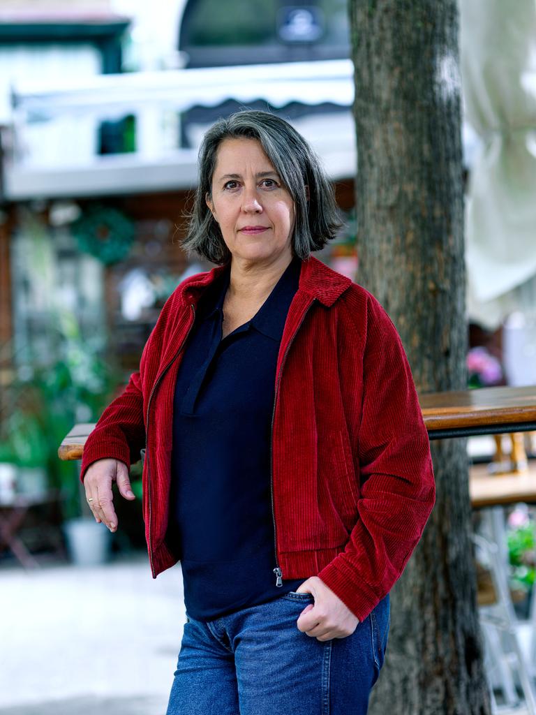 Eine Frau steht in einem Gastgarten und blickt ernst in die Kamera. Sie trägt Jeans, ein dunkelblaues Oberteil und eine rote Cordjacke. Ihre Haare sind grau bis dunkelbraun, ihre Augen braun.