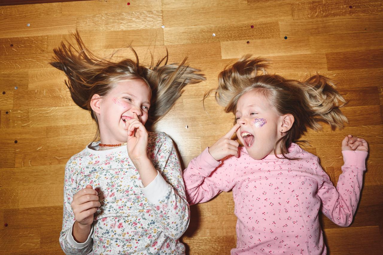Zwei Schwestern stecken den Finger in die Nase, während sie lachend auf dem Fußboden liegen.