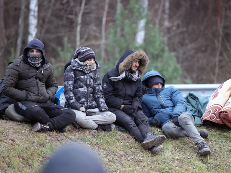 Vier Flüchtlinge, alle in dicke Winterjacken gehüllt, sitzen draußen auf dem Boden, hinter ihnen befinden sich Bäume. 