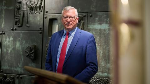 Bernhard Felmberg trägt einen blauen Anzug und rote Krawatte. Er steht vor einer Wand mit Bronzerelief und biblischen Darstellungen.