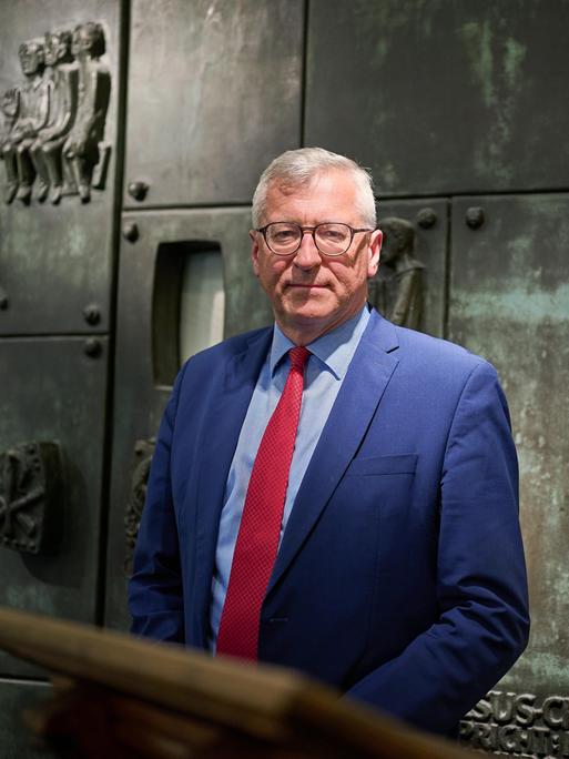 Bernhard Felmberg trägt einen blauen Anzug und rote Krawatte. Er steht vor einer Wand mit Bronzerelief und biblischen Darstellungen.