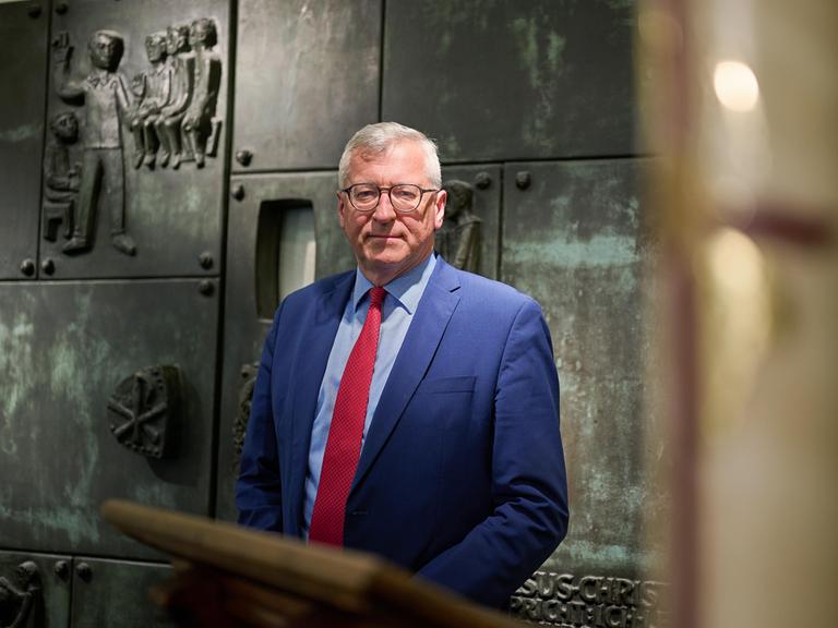 Bernhard Felmberg trägt einen blauen Anzug und rote Krawatte. Er steht vor einer Wand mit Bronzerelief und biblischen Darstellungen.