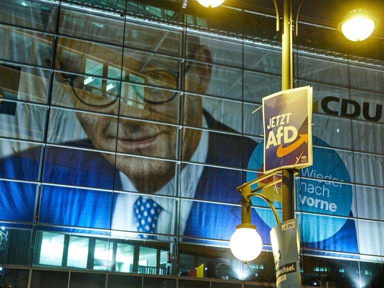 Ein Wahlplakat der AfD hängt vor der Parteizentrale der CDU, dem Konrad-Adenauer Haus. Im Hintergrund sieht man das Konterfei von Friedrich Merz auf dem Wahlplakat. 