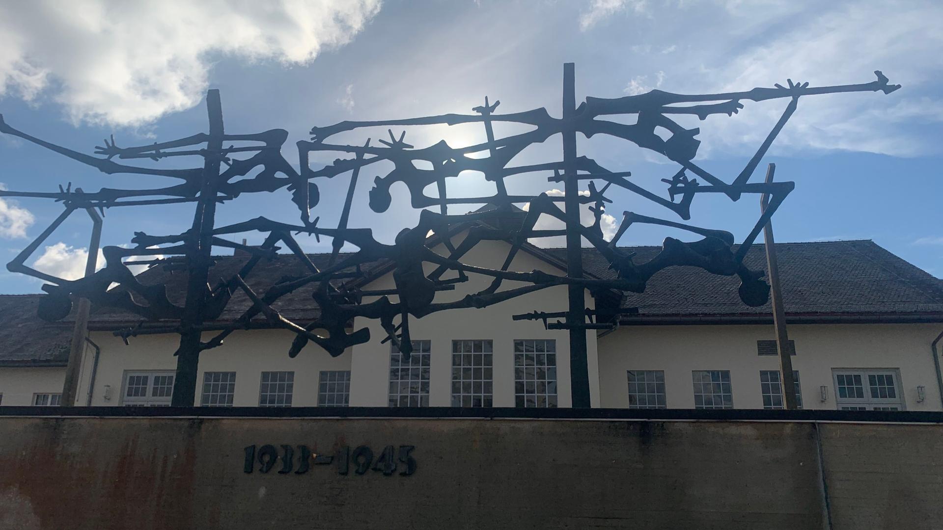Blick von unten über einen Zaun mit Stahlgeflecht und Jahreszahlen auf dem Gelände des ehemaligen KZs Dachau. Zu sehen ist eines der Gebäude, davor Eisenskulpturen mit Menschen und Stacheldrahtzaun.