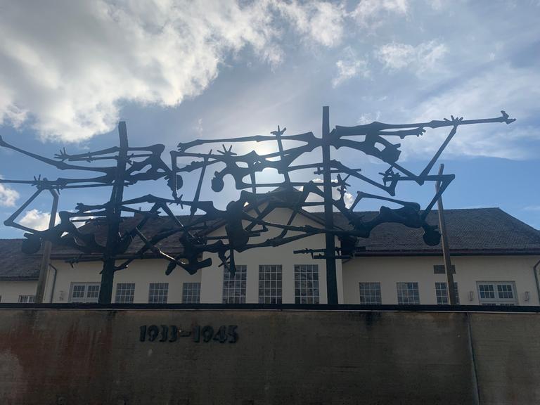 Blick von unten über einen Zaun mit Stahlgeflecht und Jahreszahlen auf dem Gelände des ehemaligen KZs Dachau. Zu sehen ist eines der Gebäude.
