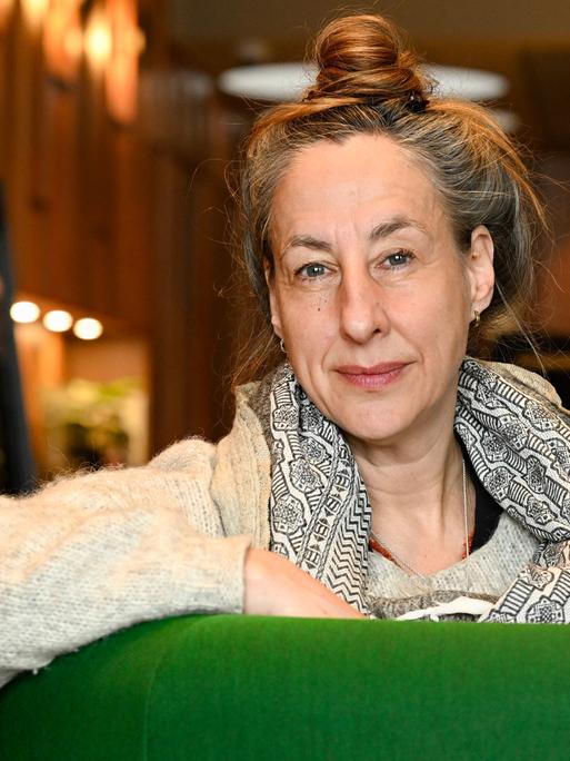 Judith Hermann lehnt über der Lehne eines grünen Sessels und blickt lächelnd in die Kamera.
