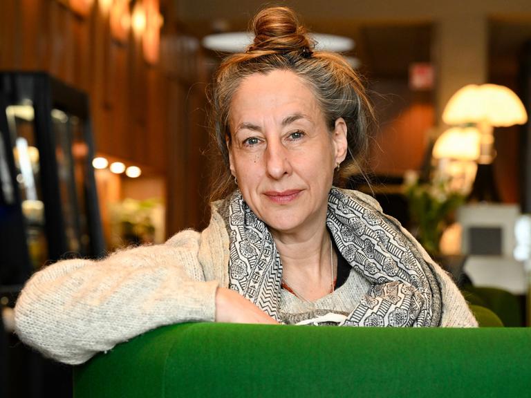 Judith Hermann lehnt über der Lehne eines grünen Sessels und blickt lächelnd in die Kamera.