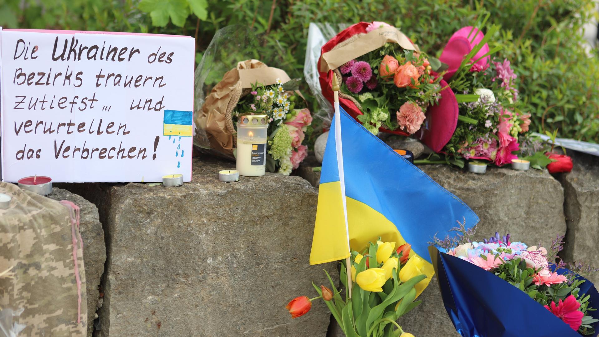 Auf einer Mauer liegen Blumen und eine ukrainische Fahne. Daneben ein Pappschild mit der Aufschrift "Die Ukrainer des Bezirks trauern zutiefst und verurteilen das Verbechen!".