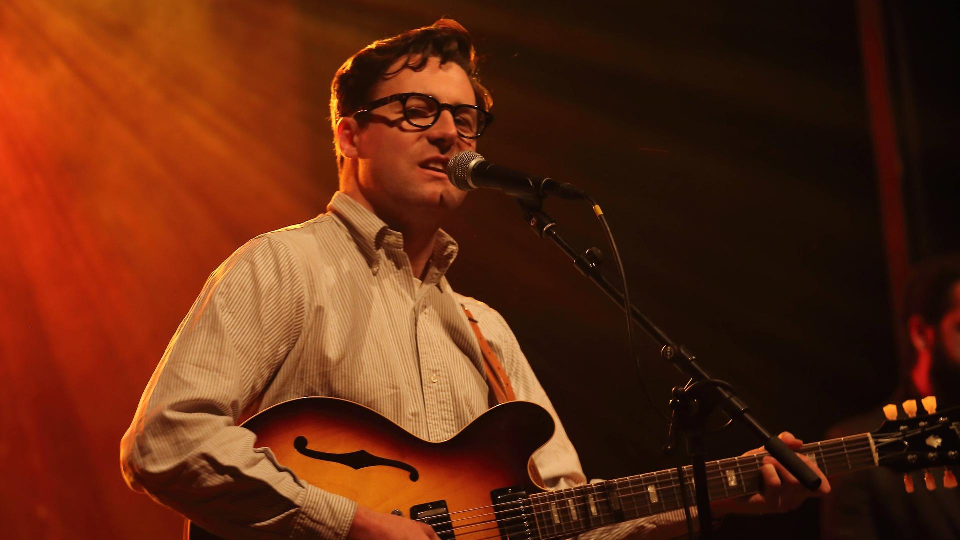 Nick Waterhouse steht mit schwarzer Brille auf einer orange ausgeleuchteten Bühne und spielt seine große Gitarre. 