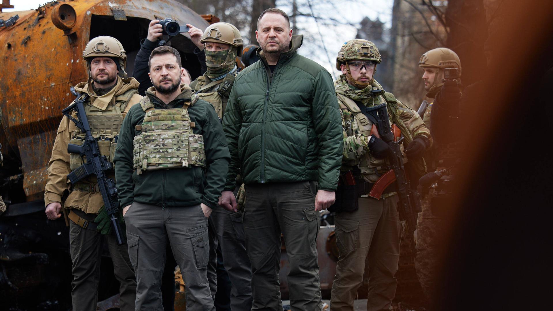 Der ukrainische Präsident Wolodymyr Selenskyj steht inmitten von Militärs und Begleitpersonen in zivil zwischen rostigen Trümmern. Der ukrainische Präsident Wolodymyr Selenskyj steht inmitten von Militärs und Begleitpersonen in zivil zwischen rostigen Trümmern.