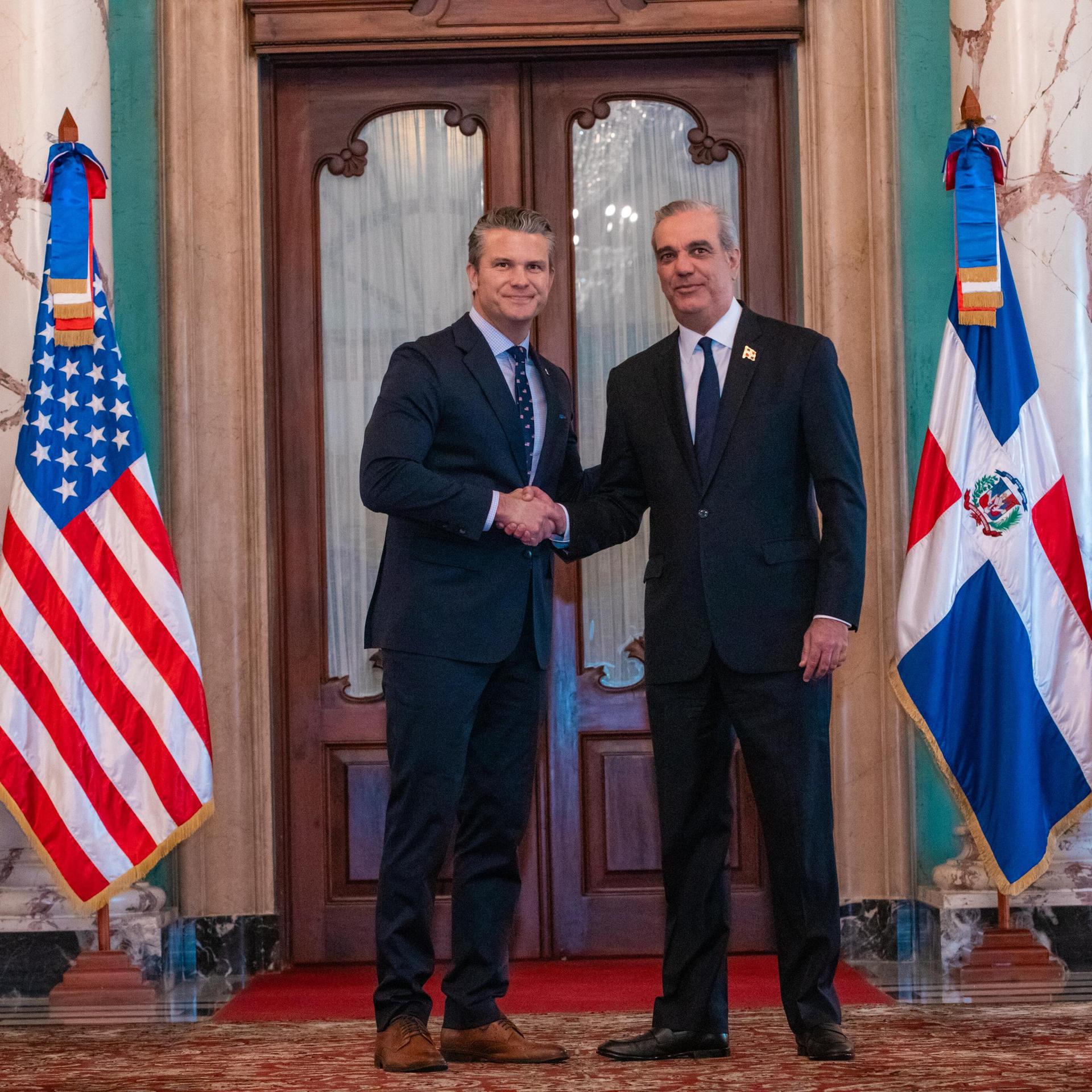 Der dominikanische Präsident Luis Abinader (rechts im Bild) schüttelt in Santo Domingo Hände mit US-Verteidigungsminister Pete Hegseth, neben den Politikern sind Flaggen beider Länder zu sehen. 