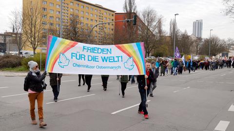 Menschen gehen auf einer Strasse in Chemnitz. Vorne wird ein Transparent von zwei Personen getragen, auf dem steht "Frieden für die Welt - Chemnitzer Ostermarsch".