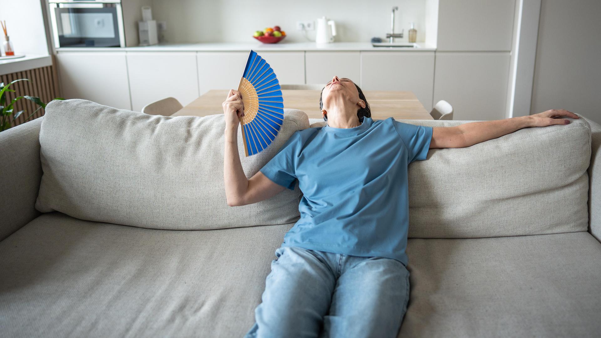 Eine Frau sitzt erschöpft auf einem Sofa in einem modernen Wohnzimmer. Sie trägt ein blaues T-Shirt und hält einen Fächer in der Hand, mit dem sie sich Luft zufächelt. Ihr Kopf ist in den Nacken gelegt, sie wirkt überhitzt und müde.