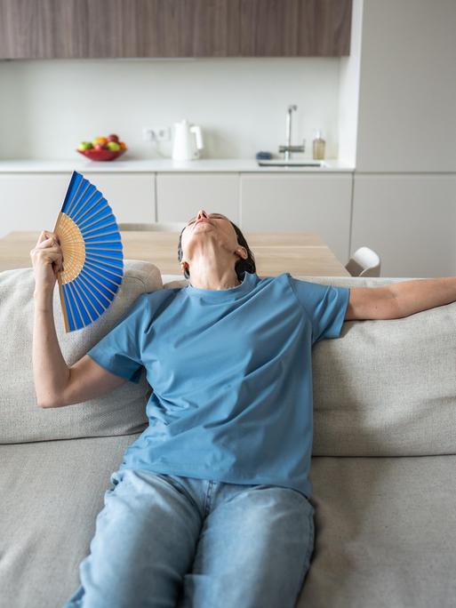 Eine Frau sitzt erschöpft auf einem Sofa in einem modernen Wohnzimmer. Sie trägt ein blaues T-Shirt und hält einen Fächer in der Hand, mit dem sie sich Luft zufächelt. Ihr Kopf ist in den Nacken gelegt, sie wirkt überhitzt und müde.