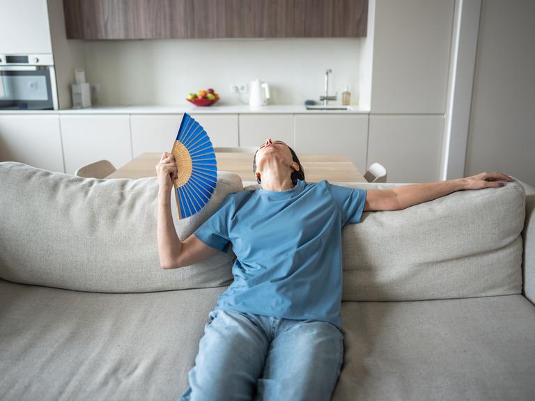 Eine Frau sitzt erschöpft auf einem Sofa in einem modernen Wohnzimmer. Sie trägt ein blaues T-Shirt und hält einen Fächer in der Hand, mit dem sie sich Luft zufächelt. Ihr Kopf ist in den Nacken gelegt, sie wirkt überhitzt und müde.