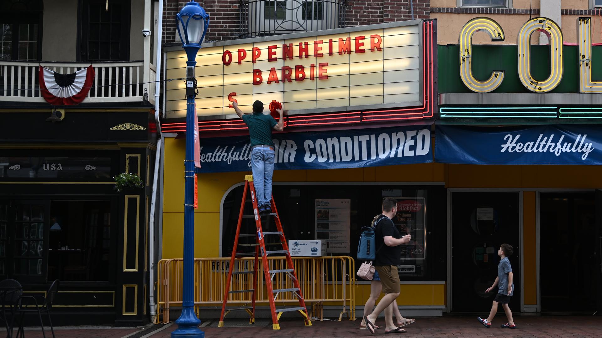 Ein Mann steht auf einer Leiter, um an der Anzeigetafel eines Kinos die beiden aktuellen Starttitel anzubringen: Barbie und Oppenheimer.