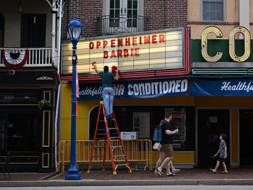 Ein Mann steht auf einer Leiter, um an der Anzeigetafel eines Kinos die beiden aktuellen Starttitel anzubringen: Barbie und Oppenheimer.