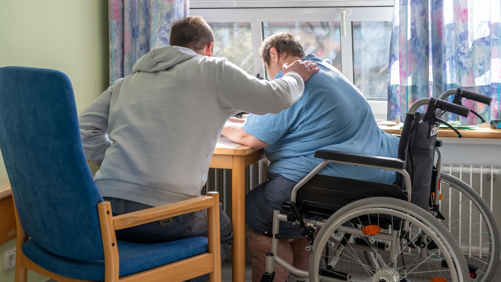 Ein Mensch legt seine Hand auf die Schulter von einer älteren Person im Roll-Stuhl. Sie sitzen zusammen an einem Tisch und sind von hinten zu sehen. Ein Mensch legt seine Hand auf die Schulter von einer älteren Person im Roll-Stuhl. Sie sitzen zusammen an einem Tisch und sind von hinten zu sehen.