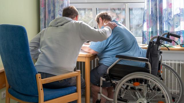 Ein Mensch legt seine Hand auf die Schulter von einer älteren Person im Roll-Stuhl. Sie sitzen zusammen an einem Tisch und sind von hinten zu sehen. 